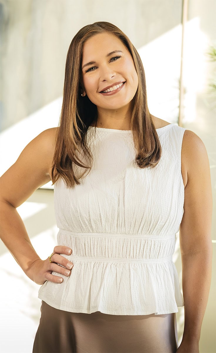 Smiling woman with long brown hair in a white sleeveless top at Arya Medical Spa.